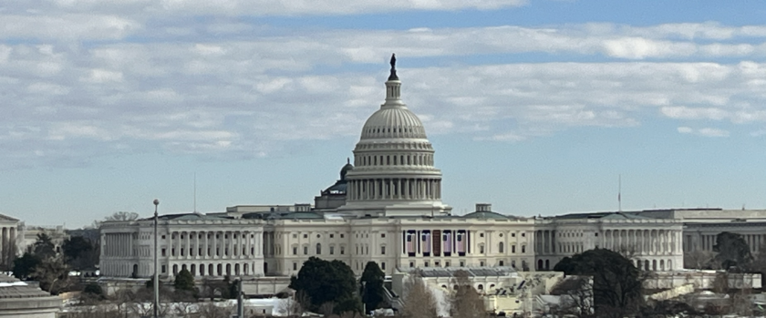 Photographic of the iconic white Capitol Building with its distinctive dome and four massive columns, set against a blue sky with white puffy clouds.